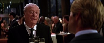 Movie still from “The Dark Knight” (2008), directed by Christopher Nolan – An older man wearing a suit and tie at an event; Close Up shot, Over the shoulder angle