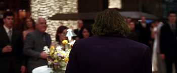 Movie still from “The Dark Knight” (2008), directed by Christopher Nolan – A man with long hair standing in front of a group of people; Medium shot, Over the shoulder angle