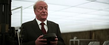 Movie still from “The Dark Knight” (2008), directed by Christopher Nolan – An older man in a suit and tie holding a remote control; Close Up shot, Over the shoulder angle