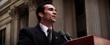 Movie still from “The Dark Knight” (2008), directed by Christopher Nolan – A man in a suit and tie speaking at a podium; Close Up shot, Low angle