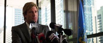 Movie still from “The Dark Knight” (2008), directed by Christopher Nolan – A man in a gray suit sitting in front of microphones and microphones; Close Up shot, Over the shoulder angle