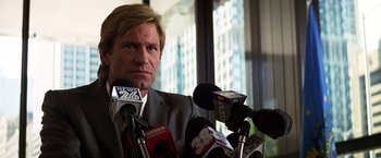 Movie still from “The Dark Knight” (2008), directed by Christopher Nolan – A man in a gray suit is sitting in front of microphones; Close Up shot, Low angle