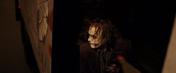 Movie still from “The Dark Knight” (2008), directed by Christopher Nolan – A man in a suit and a tie is sitting in a dark room; Medium shot, Over the shoulder angle