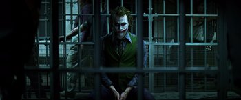 Movie still from “The Dark Knight” (2008), directed by Christopher Nolan – A man with a joker mask sitting in front of a jail cell; Medium shot, Low angle