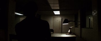 Movie still from “The Dark Knight” (2008), directed by Christopher Nolan – A man sitting at a table in a dimly - lit room; Wide shot, Low angle