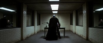Movie still from “The Dark Knight” (2008), directed by Christopher Nolan – A man sitting at a table in a room with a batman costume on; Wide shot, Low angle