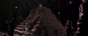 Movie still from “The Dark Knight” (2008), directed by Christopher Nolan – A man sitting on top of a large stack of cans; Wide shot, Low angle