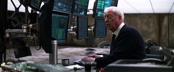 Movie still from “The Dark Knight” (2008), directed by Christopher Nolan – An older man sitting in front of multiple computer monitors; Medium shot, Over the shoulder angle