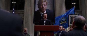Movie still from “The Dark Knight” (2008), directed by Christopher Nolan – A man is speaking at a podium in front of a portrait; Medium shot, Low angle