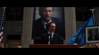 Movie still from “The Dark Knight Rises” (2012), directed by Christopher Nolan – A man in a suit and tie standing in front of a microphone; Medium shot, Low angle