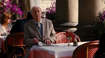 Movie still from “The Dark Knight Rises” (2012), directed by Christopher Nolan – An older man sitting at a table in front of flowers; Medium shot, Over the shoulder angle