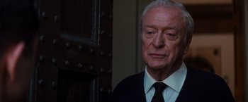 Movie still from “The Dark Knight Rises” (2012), directed by Christopher Nolan – An older man wearing a suit and tie standing in front of a wooden door; Close Up shot, Over the shoulder angle
