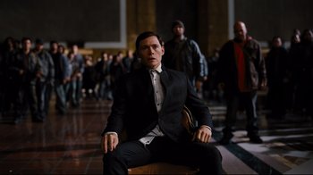 Movie still from “The Dark Knight Rises” (2012), directed by Christopher Nolan – A man in a suit and tie sitting in a chair in front of a group of people; Medium shot, High angle