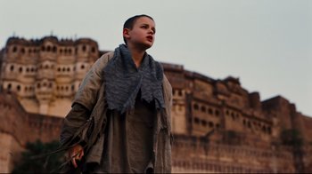 Movie still from “The Dark Knight Rises” (2012), directed by Christopher Nolan – A young boy wearing a scarf standing in front of an old building; Medium shot, Low angle