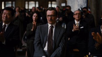 Movie still from “The Dark Knight Rises” (2012), directed by Christopher Nolan – A man in a suit sitting in front of a group of people; Medium shot, Low angle