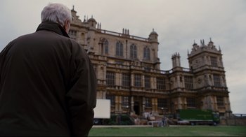Movie still from “The Dark Knight Rises” (2012), directed by Christopher Nolan – An older man standing in front of a large building; Wide shot, Over the shoulder angle