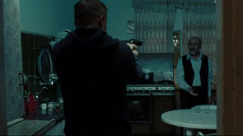 Movie still from “The Hurt Locker” (2008), directed by Kathryn Bigelow – A man holding a gun in a kitchen while a woman stands in front of a stove; Medium shot, Over the shoulder angle