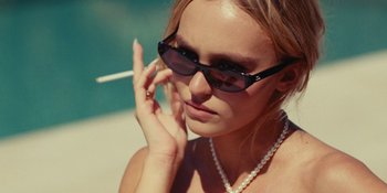 Movie still from “The Idol” (2023), directed by Sam Levinson – A beautiful woman wearing sunglasses and a pearl necklace smoking a cigarette while talking on a cell phone; Close Up shot, Low angle
