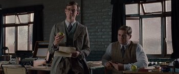 Movie still from “The Imitation Game” (2014), directed by Morten Tyldum – A man in a suit and tie holding an apple and standing next to another man in a suit and tie; Medium shot, Low angle