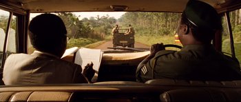 Movie still from “The Last King of Scotland” (2006), directed by Kevin Macdonald – A view from the back seat of a car of a truck driving down a road; Medium shot, Over the shoulder angle