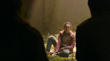 Movie still from “The Last of Us” (2023), created by Neil Druckmann – A woman sitting on the ground in the woods; Wide shot, High angle