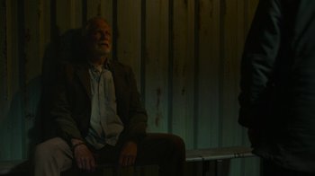 Movie still from “The Last of Us” (2023), created by Neil Druckmann – An older man sitting in a chair in a dark room; Medium shot, Low angle