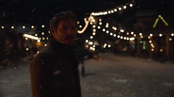 Movie still from “The Last of Us” (2023), created by Neil Druckmann – A man standing in the snow at night with lights in the background; Medium shot, Over the shoulder angle