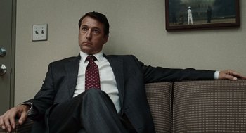Movie still from “Burn After Reading” (2008), directed by Joel Coen – A man in a suit and tie sitting on a couch; Medium shot, Low angle