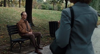 Movie still from “Burn After Reading” (2008), directed by Joel Coen – An older man sitting on a park bench while taking a picture; Medium shot, Over the shoulder angle
