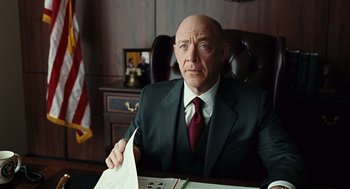 Movie still from “Burn After Reading” (2008), directed by Joel Coen – A man in a suit and tie sitting at a desk holding papers; Medium shot, Over the shoulder angle