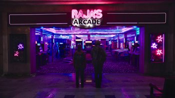 Movie still from “The Last of Us” (2023), created by Neil Druckmann – Two people standing in front of the entrance to rajas arcade; Extreme Wide shot, High angle