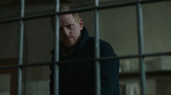 Movie still from “The Last of Us” (2023), created by Neil Druckmann – A man standing behind bars in a jail cell; Medium shot, Over the shoulder angle