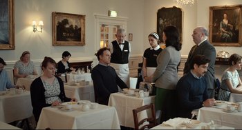 Movie still from “The Lobster” (2015), directed by Yorgos Lanthimos – A group of people standing around a table with food on it; Medium shot, Over the shoulder angle