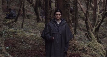 Movie still from “The Lobster” (2015), directed by Yorgos Lanthimos – A woman in a black raincoat standing next to a tree in the woods; Medium shot, Over the shoulder angle