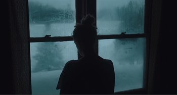 Movie still from “The Lodge” (2019), directed by Severin Fiala – A woman sitting in front of a window looking outside; Medium shot, Over the shoulder angle