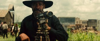 Movie still from “The Magnificent Seven” (2016), directed by Antoine Fuqua – A man in a black shirt and a black hat holding a camera; Medium shot, Low angle