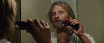 A man with long hair and a goatee shaving in a bathroom mirror