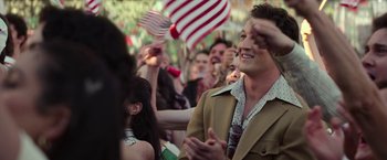 Movie still from “The Offer” (2022), directed by Dexter Fletcher – A man in a crowd of people with a flag in the background; Medium shot, Over the shoulder angle