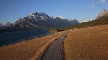 Movie still from “The Shining” (1980), directed by Stanley Kubrick – A car driving down a road near a body of water and mountains; Extreme Wide shot, High angle
