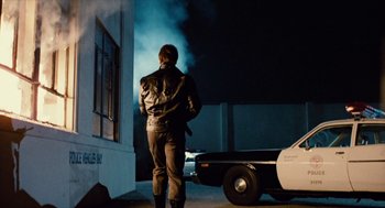 Movie still from “The Terminator” (1984), directed by James Cameron – A man standing next to a police car at night; Wide shot, Over the shoulder angle