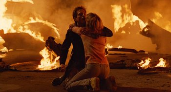 Movie still from “The Terminator” (1984), directed by James Cameron – A man and a woman hugging in front of a fire; Medium shot, High angle