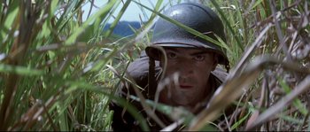 Movie still from “The Thin Red Line” (1998), directed by Terrence Malick – A man wearing a helmet is hiding in the grass; Close Up shot, Low angle