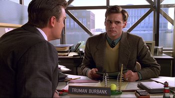 Movie still from “The Truman Show” (1998), directed by Peter Weir – Two men are sitting at a desk in front of a sign; Medium shot, Over the shoulder angle
