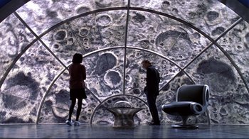 Movie still from “The Truman Show” (1998), directed by Peter Weir – A man and a woman standing in front of an image of the moon; Wide shot, High angle