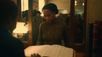 Movie still from “The Underground Railroad” (2021), directed by Barry Jenkins – A woman in a brown dress looking at a sheet of music; Medium shot, Over the shoulder angle