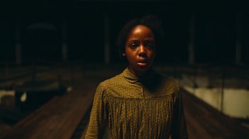 Movie still from “The Underground Railroad” (2021), directed by Barry Jenkins – A woman in a green dress standing in a dimly - lit room; Medium shot, Low angle