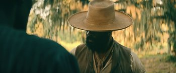 Movie still from “The Underground Railroad” (2021), directed by Barry Jenkins – A man with a beard wearing a straw hat; Close Up shot, Over the shoulder angle