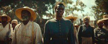 Movie still from “The Underground Railroad” (2021), directed by Barry Jenkins – A woman in a green dress standing in front of a group of people; Medium shot, Over the shoulder angle