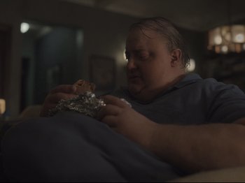 Movie still from “The Whale” (2022), directed by Darren Aronofsky – A man sitting on a couch eating a piece of food; Medium shot, High angle
