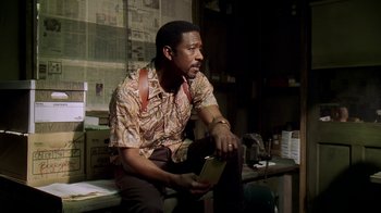 Movie still from “The Wire” (2002), created by David Simon – A man sitting on a table holding a book; Medium shot, Low angle